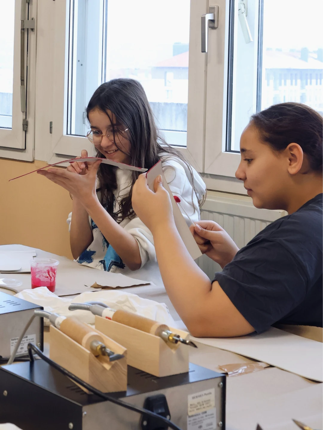 Deux élèves observant avec attention leurs pièces de cuir durant la fabrication d'un objet, avec des fers à fileter électriques posés au premier plan.